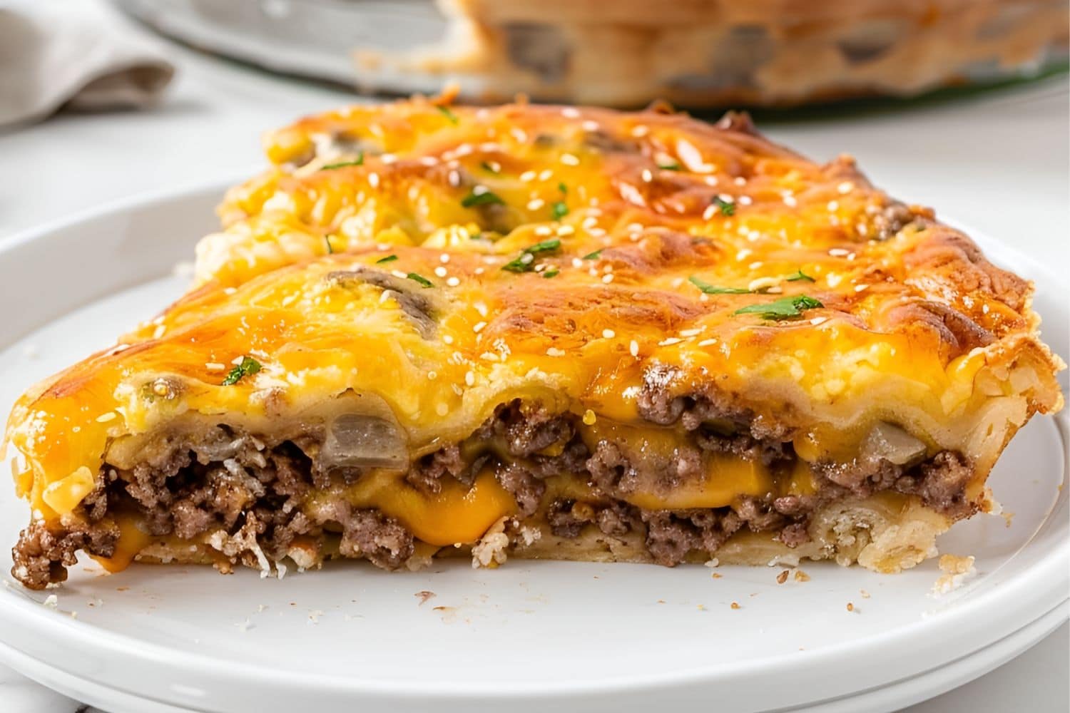 Close Up of Slice of Bisquick Cheeseburger Pie with Lots of Cheese, Ground Beef, and Onions on a White Plate with the Remaining Pie in the Dish in the Background