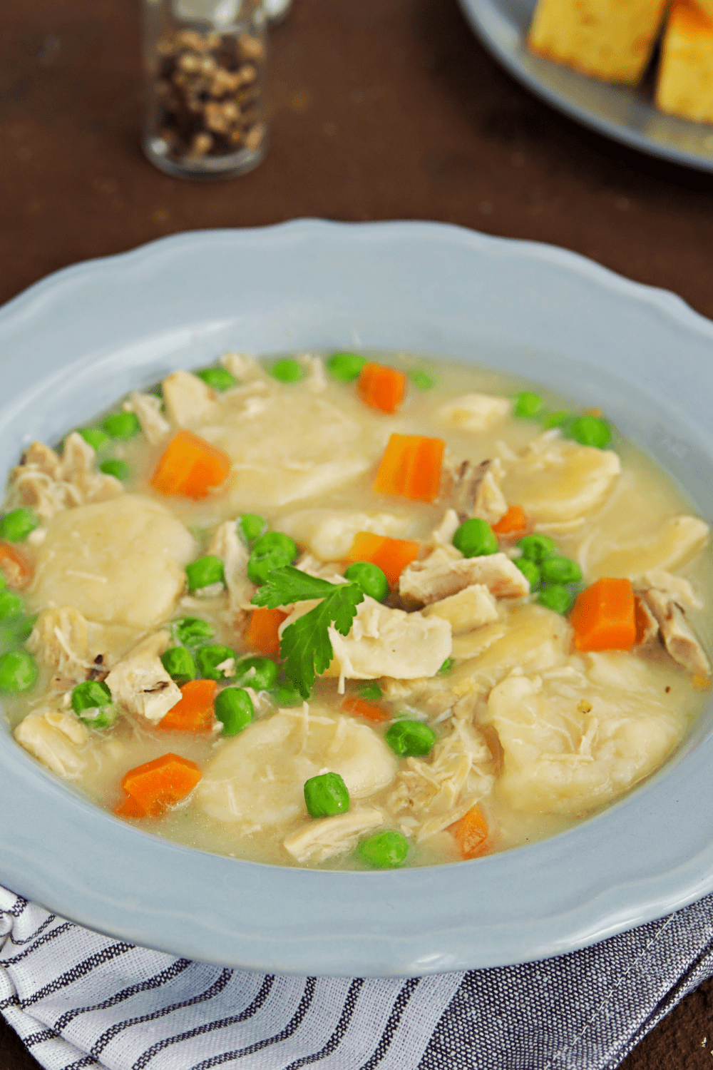 A bowl of chicken and dumplings with chopped carrots, peas, and fresh parsley on top.