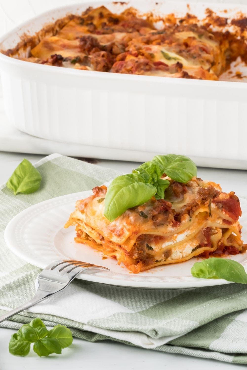 Slice of Homemade Ragu Lasagna with 3 layers of Noodles, Cheese Sauce, and Meat Sauce on a Plate with a Basil Garnish and Basil All Around