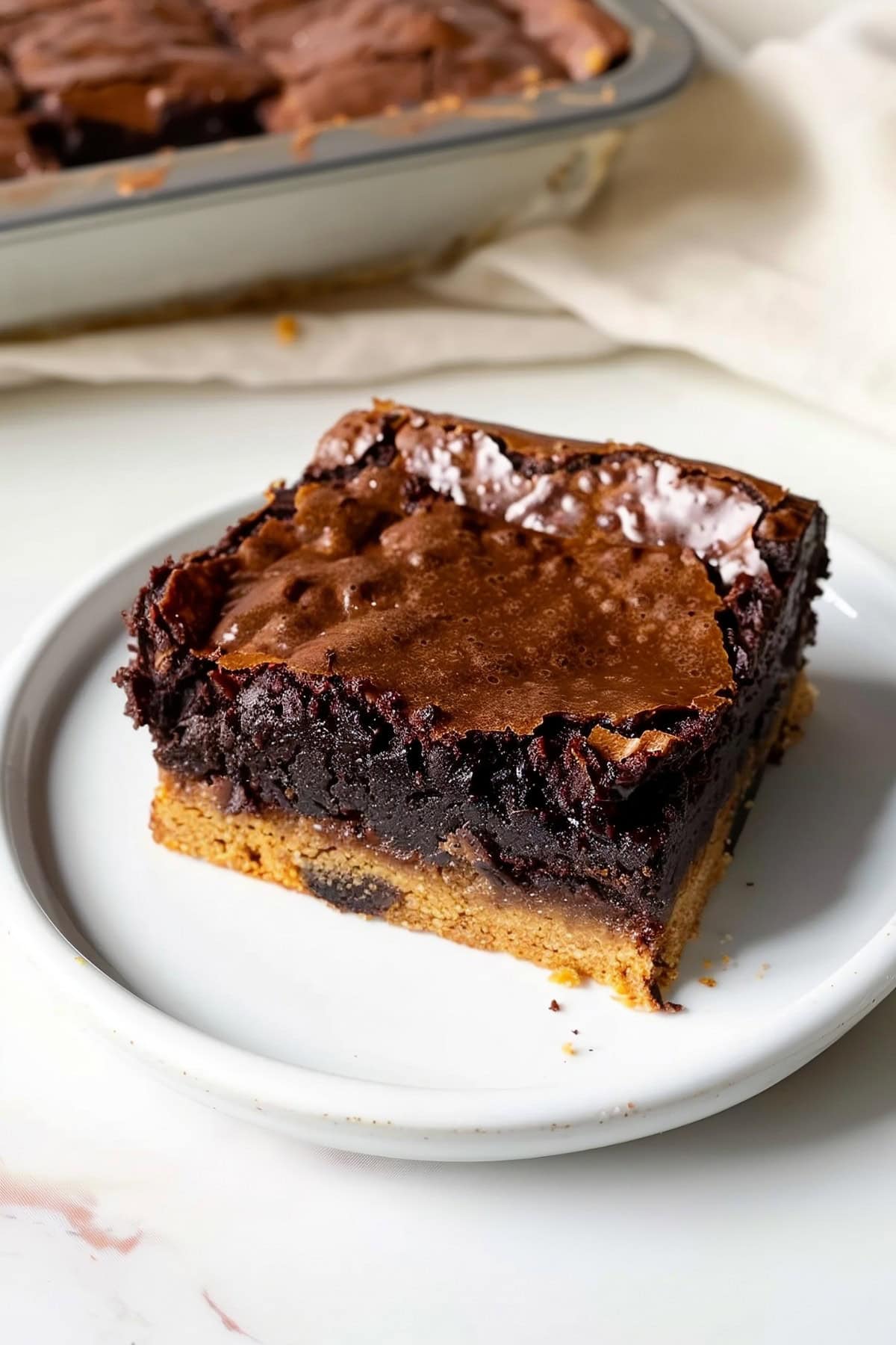 Delicious Brookie with a Brownie Top and Chocolate Chip Cookie Bottom on a White Plate with a Tray of Brookies in the Background