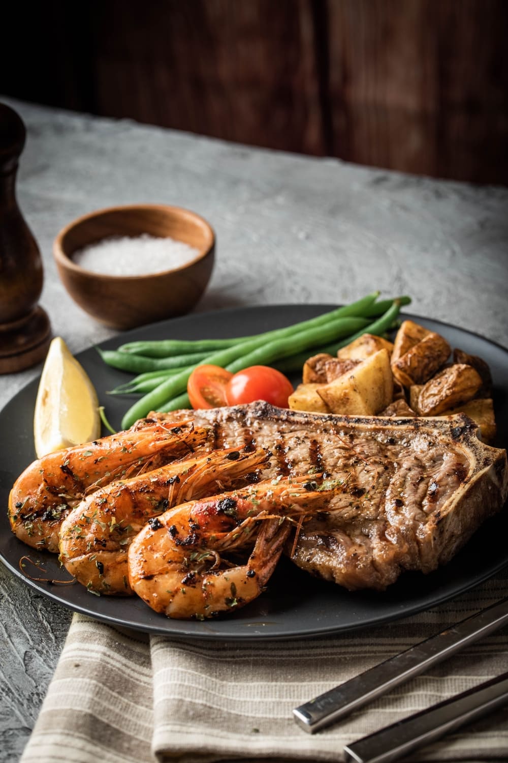 Surf and Turf with Shrimp and Strip Steak Served on a Black Plate With Roasted Potatoes, Sliced Lemon and Tomatoes and Greens on the side