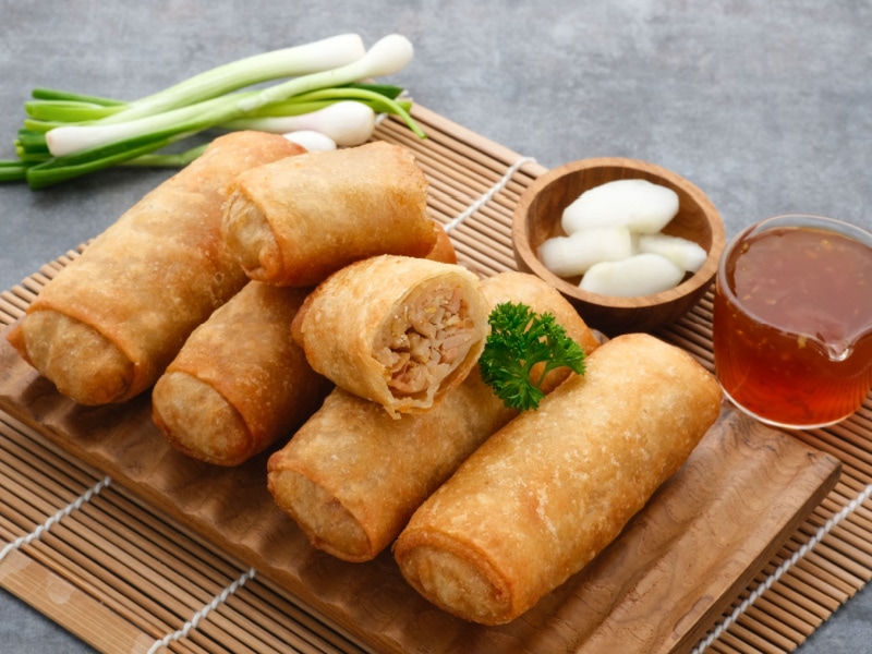 Fried egg rolls with dipping sauce and pickled garlic served on a wooden board on top of bamboo mat. 