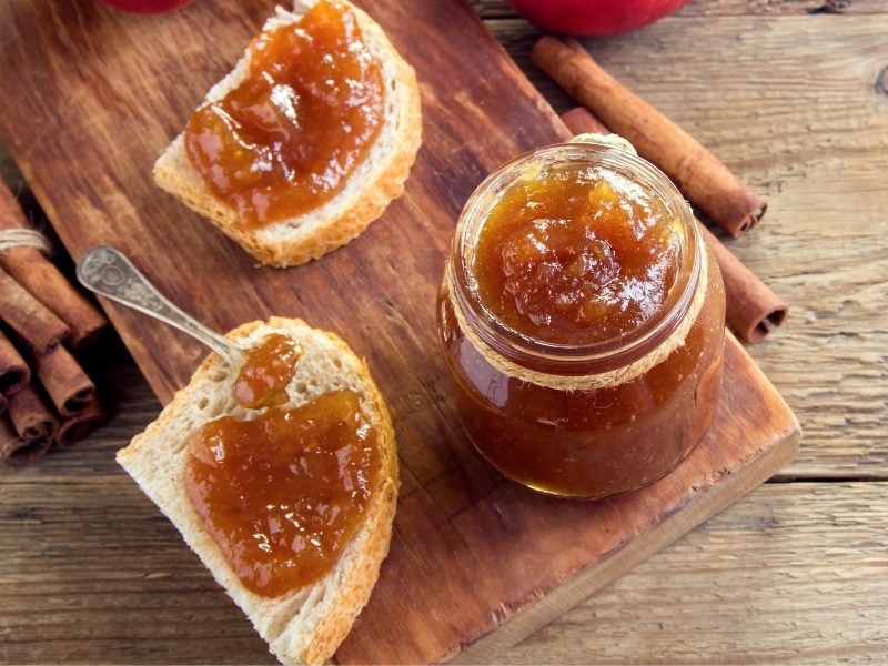 Top view of jar of apple butter, cinnamon sticks and slices of bread with spread. 