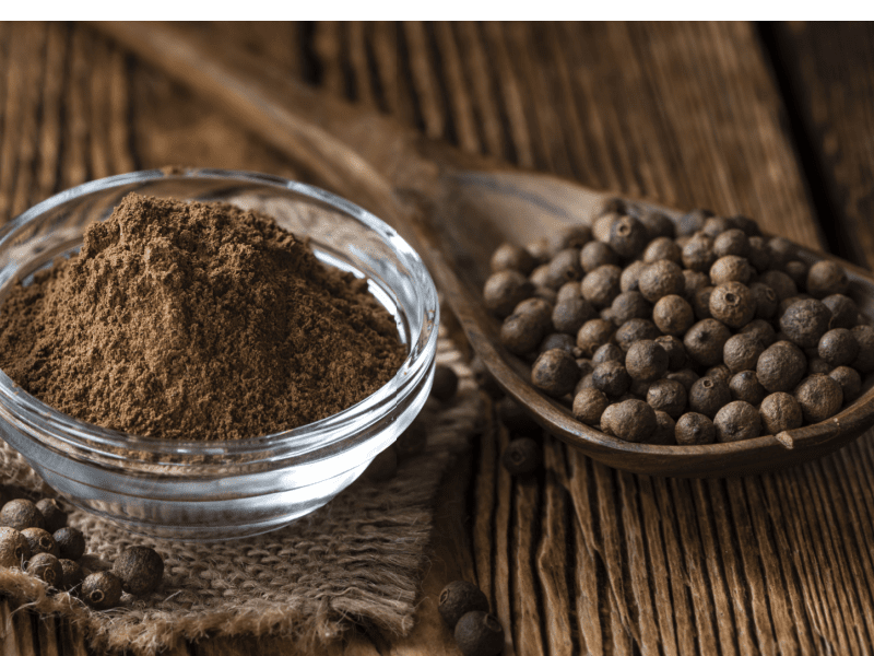 Ground Allspice in a Clear Glass Spice Dish, Whole Allspice on a Wooden Spoon Placed on Top of Wooden Table