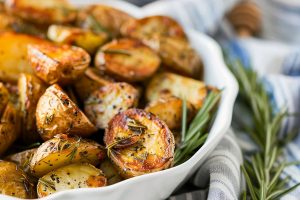Close Up of Rosemary Roasted Potatoes in a White Serving Bowl with a Striped Kitchen Towel and Sprigs of Fresh Rosemary