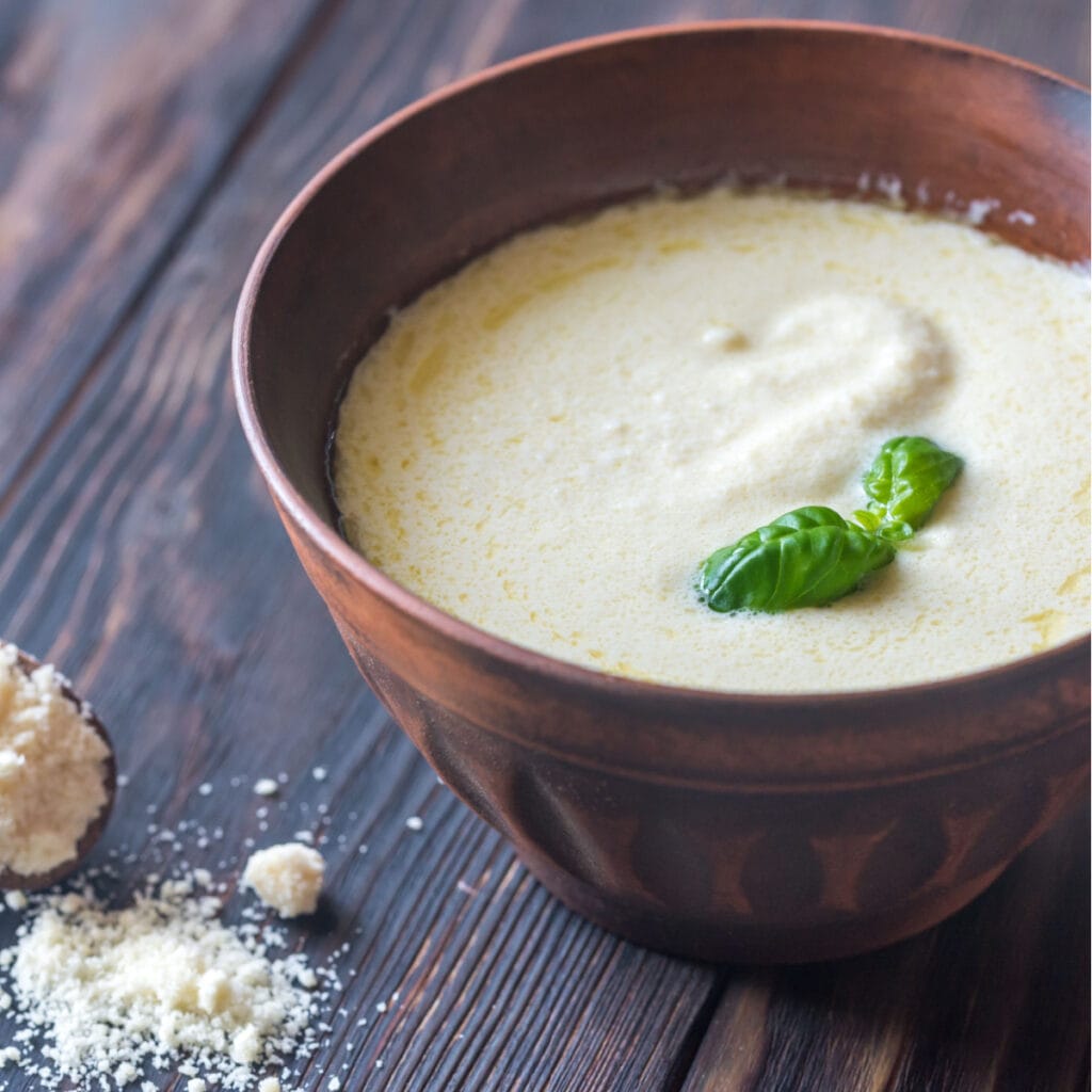 Bowl of Homemade Alfredo Sauce with Basil on a Wooden Table, Parmesan Cheese Spilled in Bottom Left Corner