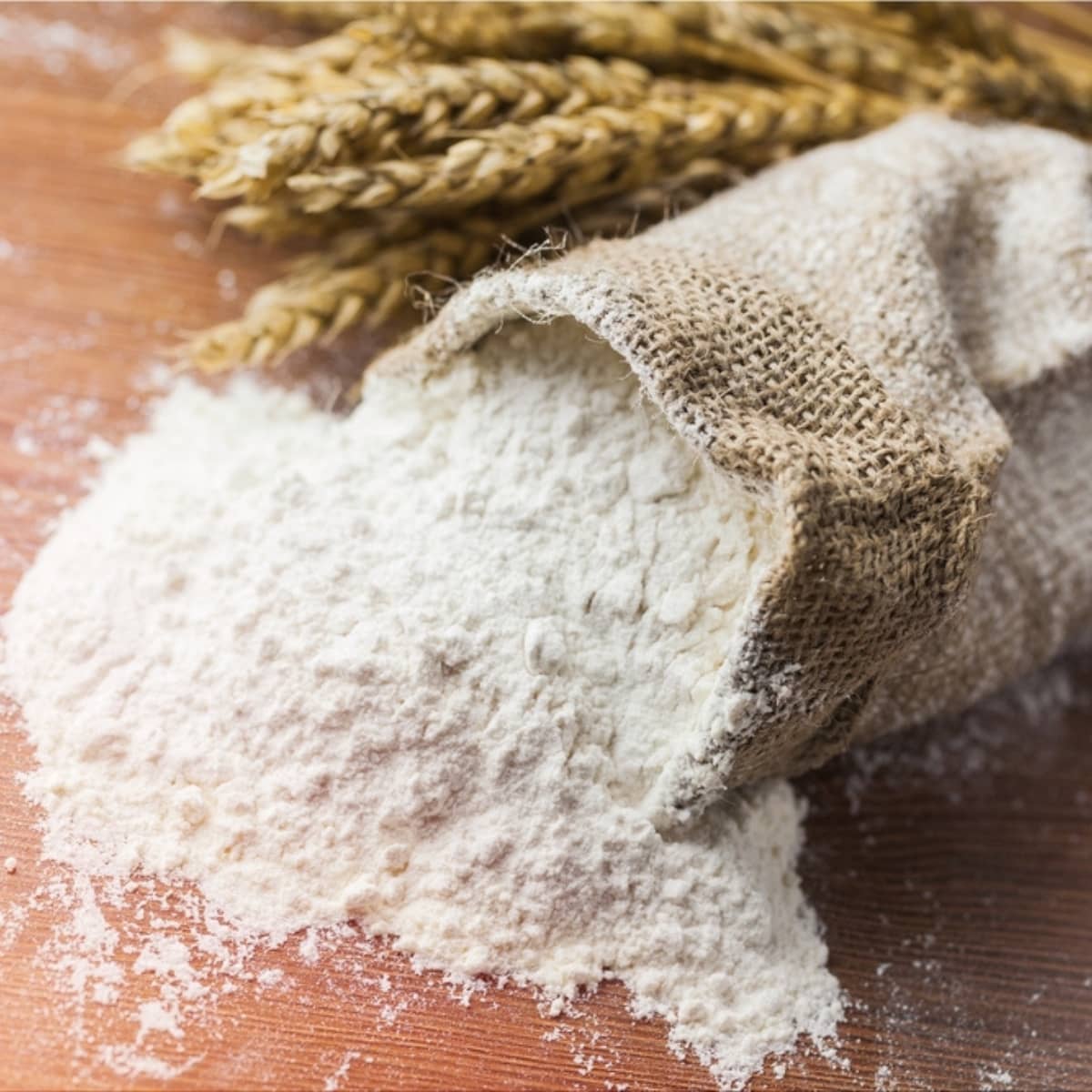 Cake Flour vs. All-Purpose Flour (What's the Difference?) featuring Burlap Bag With Flour Pouring on the Table and Stalks of Wheat in the Background