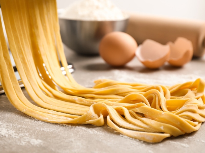 Homemade Egg Noodles Hanging from the Pasta Maker with An Egg, A Cracked Egg, a Bowl of Flour, and A Rolling Pin in the Background