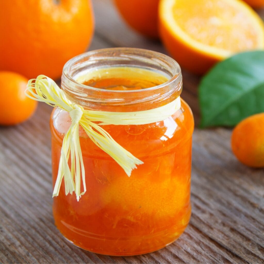 Jar of Orange Marmalade With Rustic Ribbon Tied Around the Rim and Fresh Oranges in Background