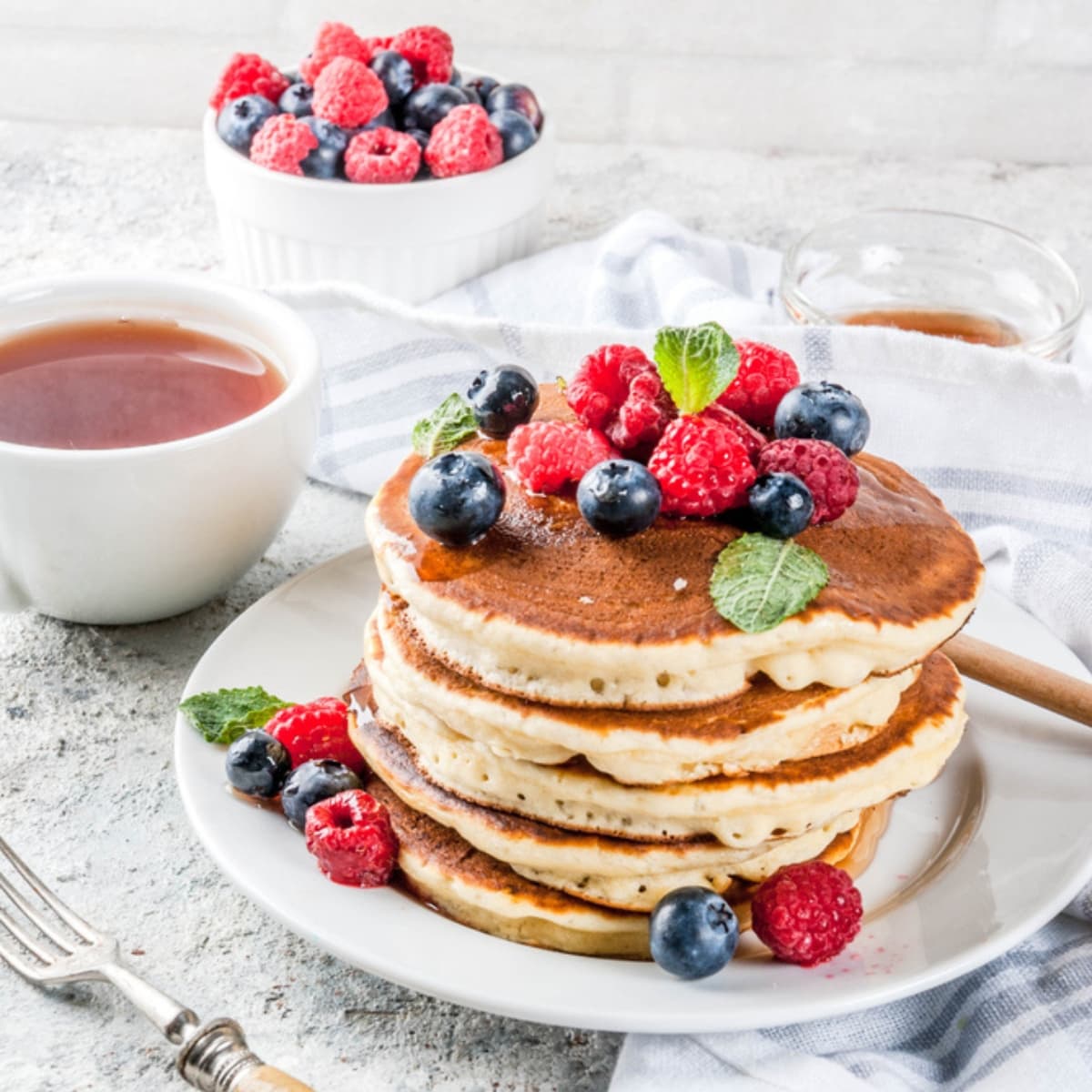 Homemade oat flour pancakes with fresh berries and mint served on a plate, dish of syrup on the side.