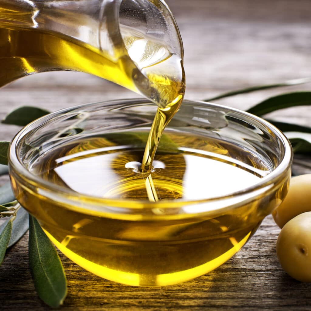 A Bottle of Olive Oil Pouring on a Bowl and Fresh Olives on a Wooden Table