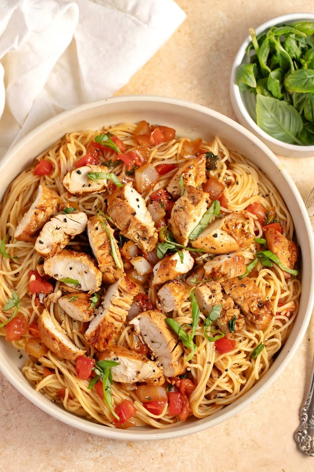 Bruschetta pasta with tomatoes, garlic, spinach, balsamic vinegar, and olive oil served on a round plate with a saucer of fresh basil leaves on the side