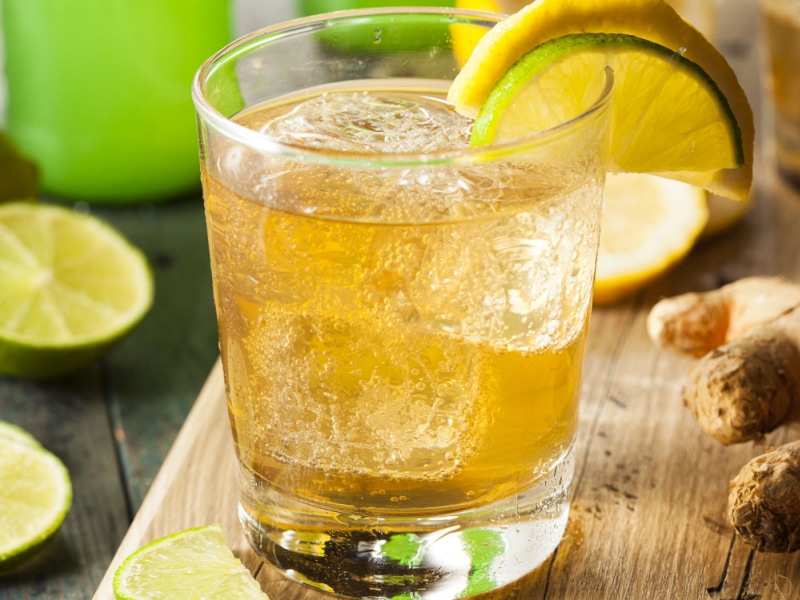 A Glass of Ginger Ale and Ice Garnished with Lime and Lemon on a Wooden Table with Lime Slices and Fresh Ginger in the Background