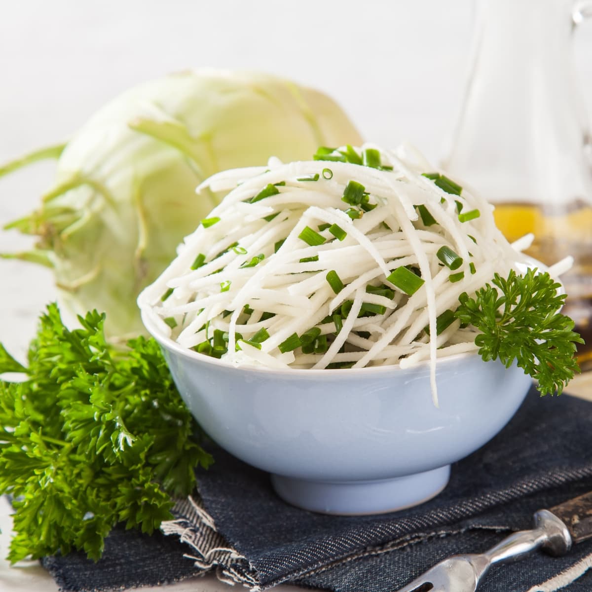 Julienned Kohlrabi in a Salad with Green Onions in a Small Blue Bowl on a Table with Silverware, Denim Cloth, Parsley, and a Whole Kohlrabi