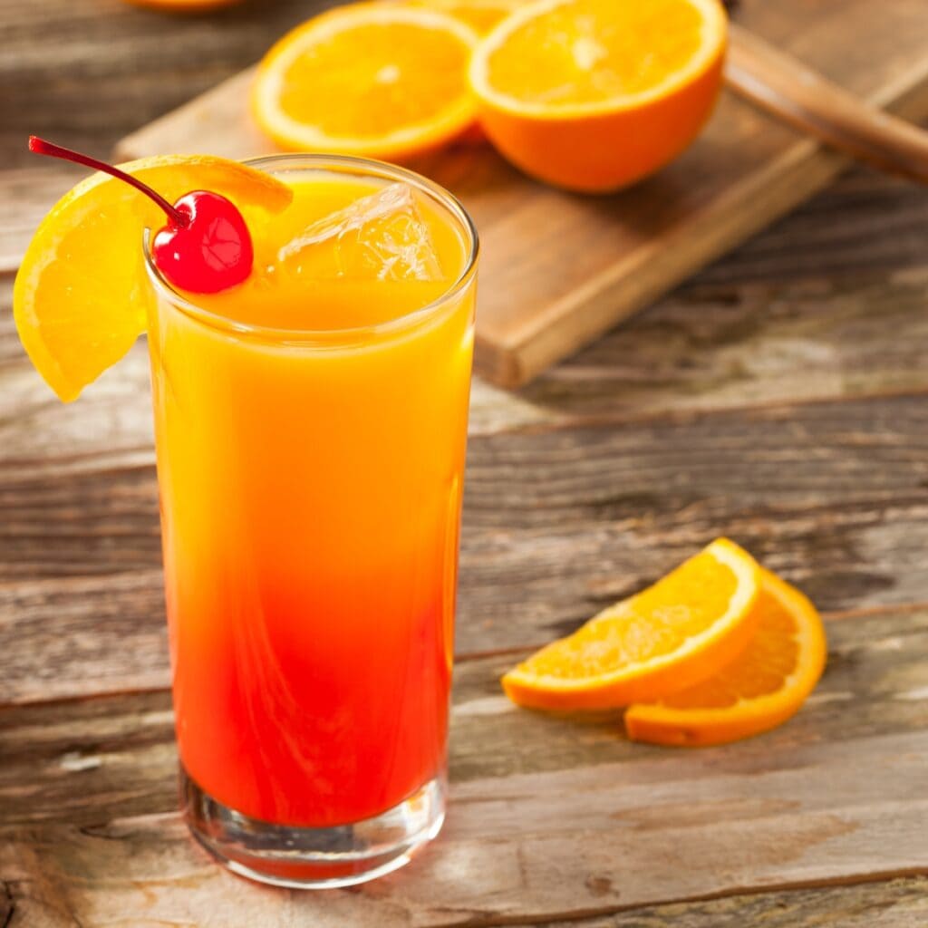 Glass of Sunrise Tequila Garnished With Orange Slice and Cherry with Halved Oranges on a Cutting Board in the Background