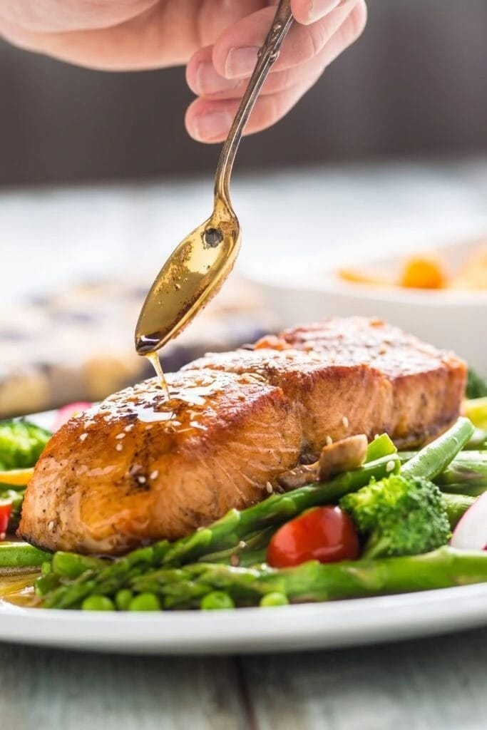Spoon pouring oil on an Air Fryer Salmon served with asparagus, broccoli and tomato