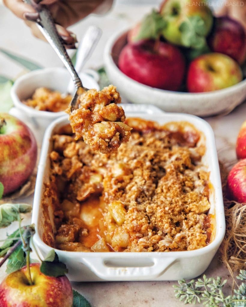 Spoon scooping a portion of vegan apple crumble from a white dish. 