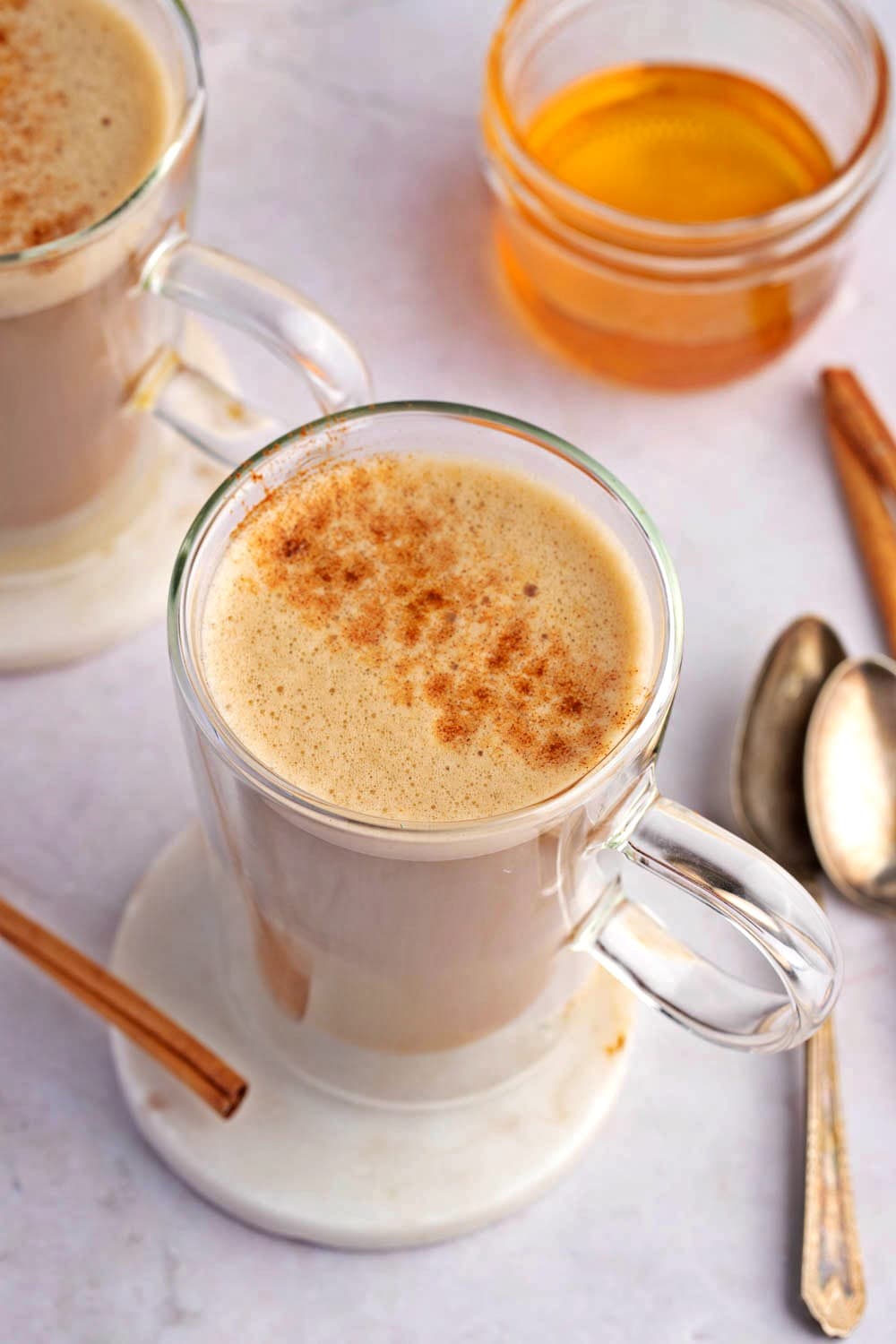 Two Clear Mugs of Warm Honey Coffee with Cinnamon and a Jar of Honey in the Background with Two Spoons and Cinnamon Sticks on the Table