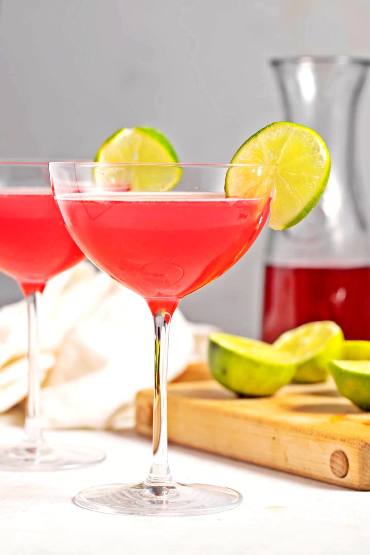 Two glasses of cosmopolitan cocktail with lime and more limes and a pitcher of cranberry juice in the background 