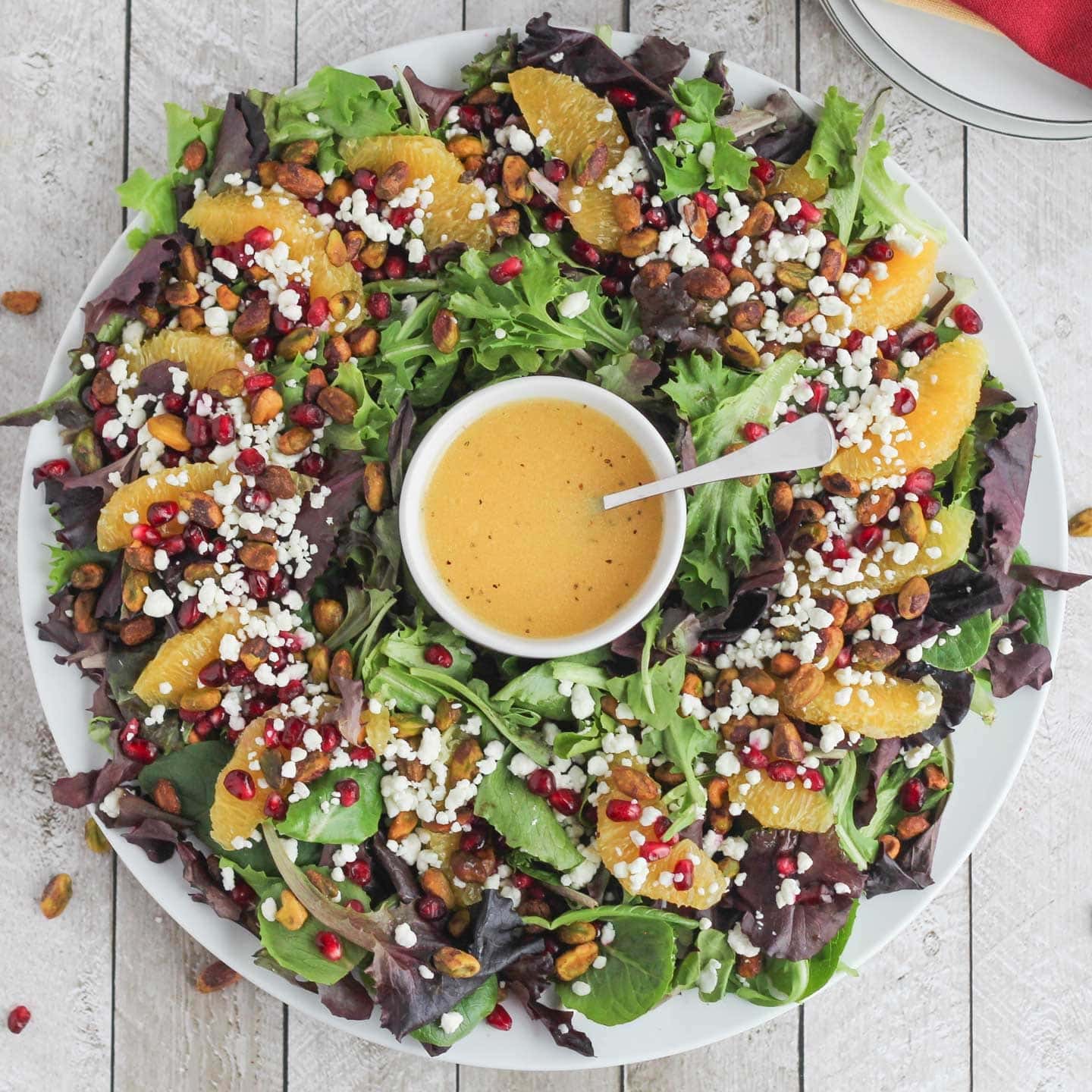 Salad with greens, pomegranate arils, goat cheese, pistachios, and oranges on a round plate. 