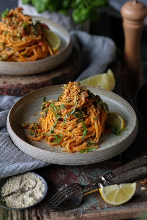 Harissa pasta with creamy sauce served on a plate with parmesan cheese, lemon slice and chopped leaves.