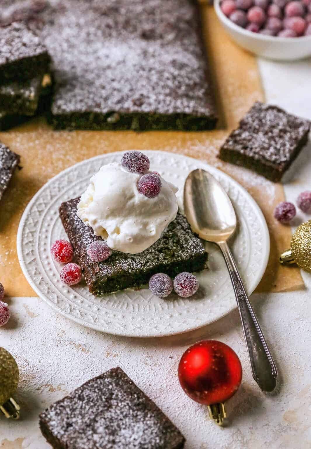 A serving of gingerbread brownie on a white plate topped with a scoop of vanilla ice cream and sugar coated berries garnish. 