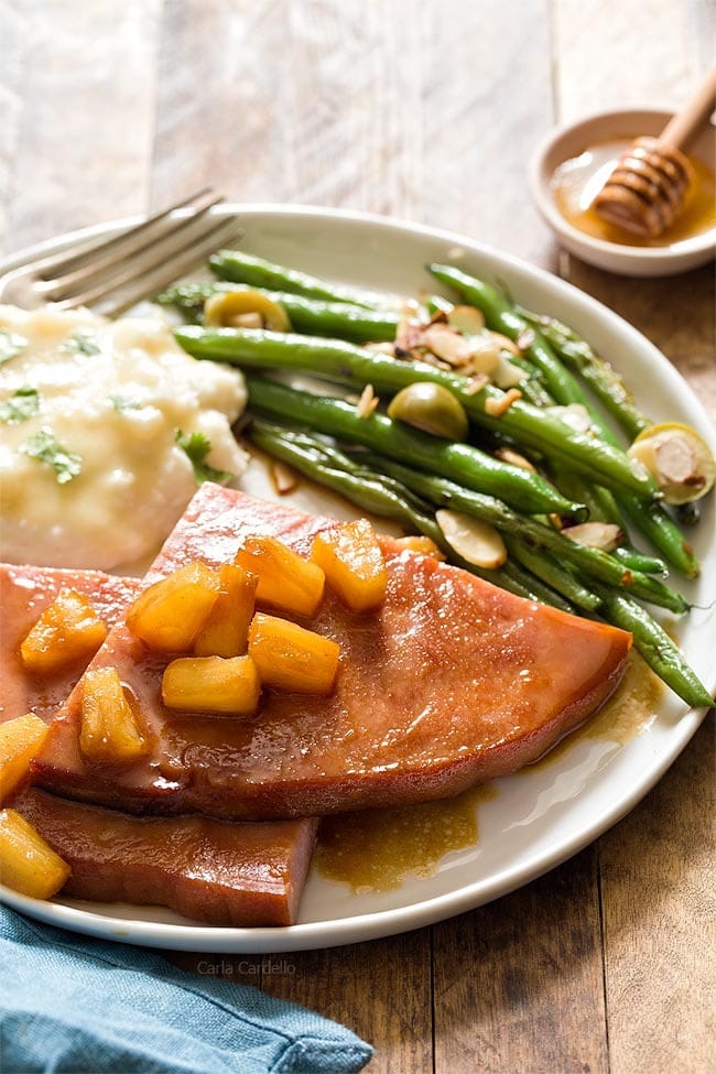 Big slices of ham served with mashed potatoes, greens garnished with pineapple chunks and honey glaze.