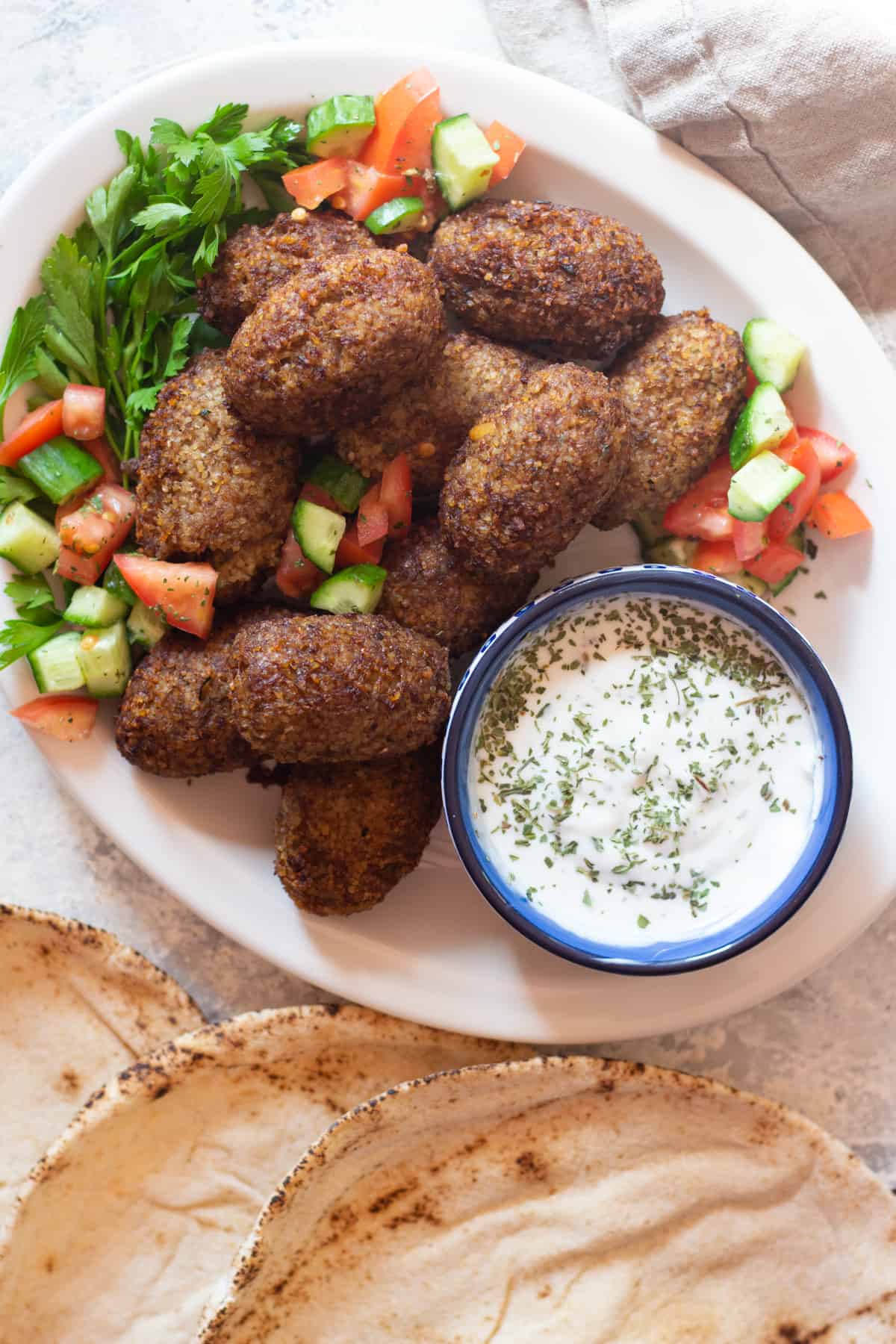 Middle Eastern Style Meatballs on a Platter with Chopped Cucumbers and Tomatoes, and a Bowl of Seasoned Yogurt Sauce and Flatbread on the Sides