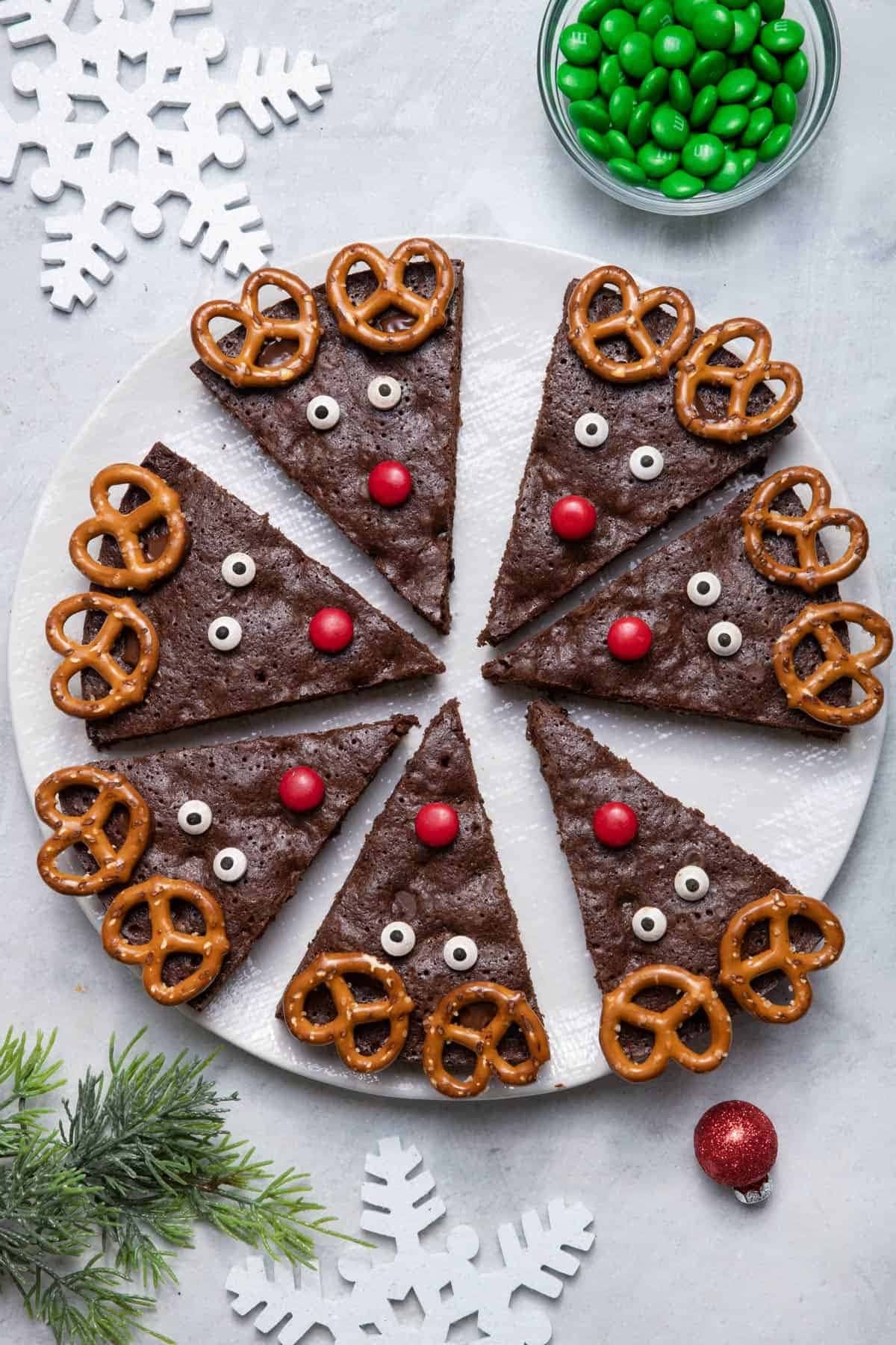 Brownies cut in triangles on a white plate decorated like reindeers with pretzels and candies. 