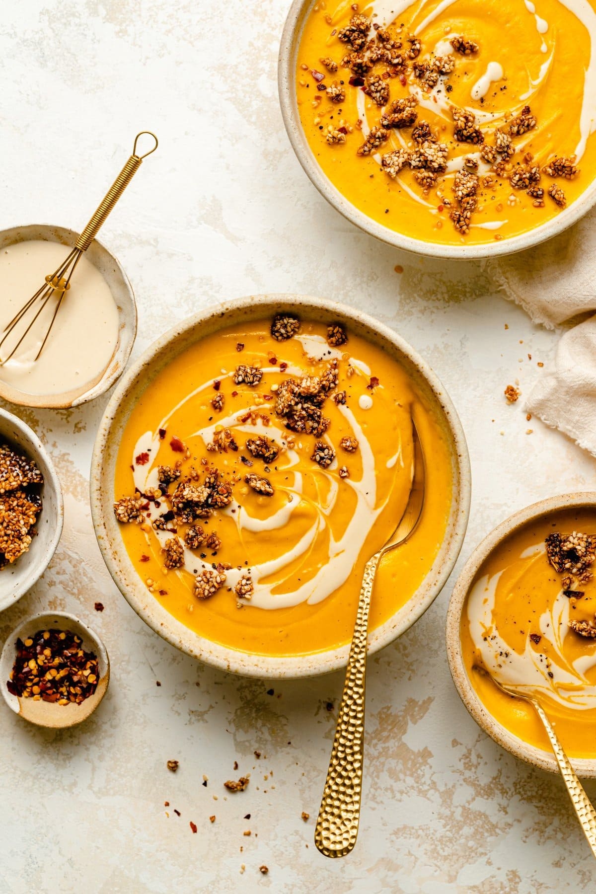 Spicy sweet potato soup with carrots, tahini, red chili flakes, and sesame brittle in two bowls with spoons