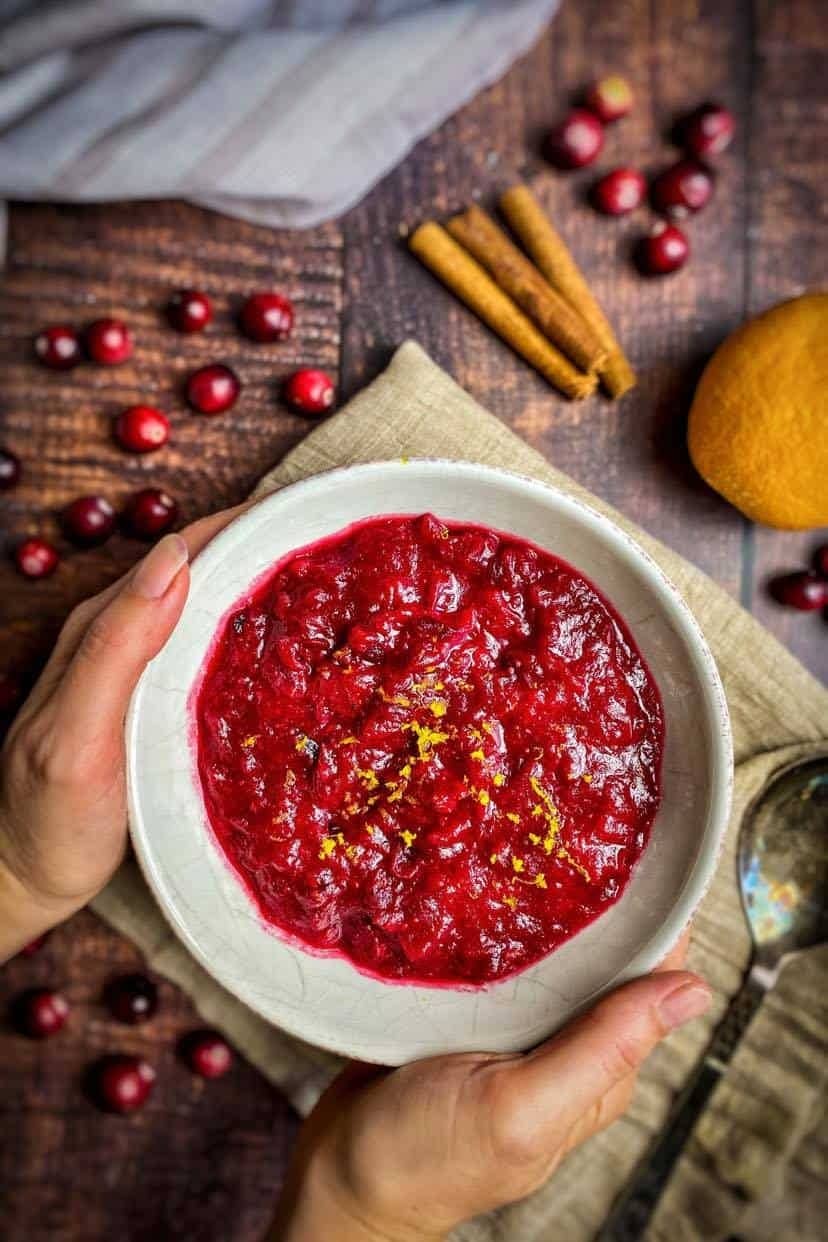 Hand holding cranberry sauce in a bowl with orange zest garnish on top. 