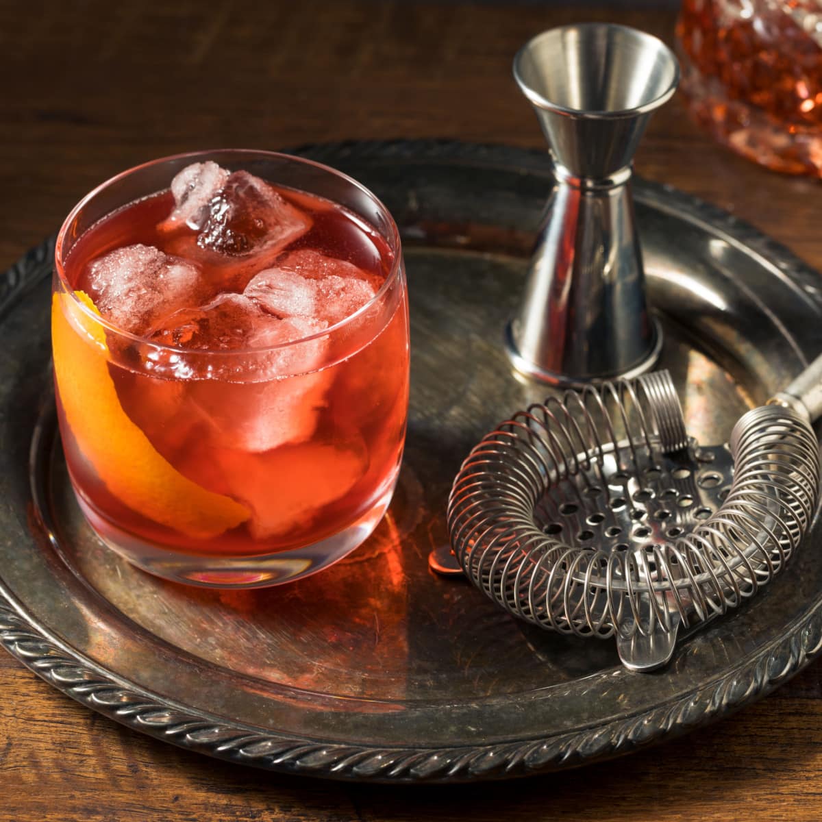 Boulevardier cocktail on glass served with ice on a steel try with cocktail jigger and strainer. 