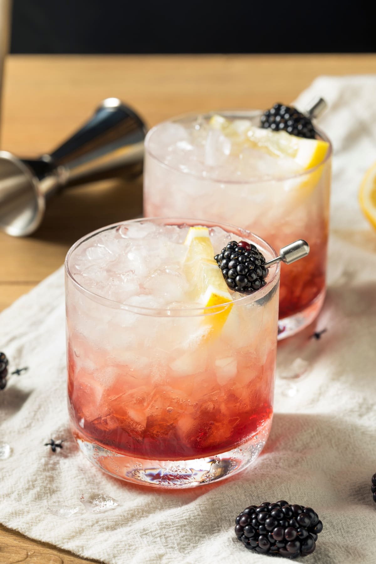 Two glasses of Bramble cocktail filled with crushed ice garnished with blackberries on skewers and slice of lemon on top of a cloth.