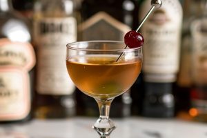 Side View Brooklyn Cocktail in a Coupe Glass with Luxardo Maraschino Cherry and Bottles of Liqueurs in the Background