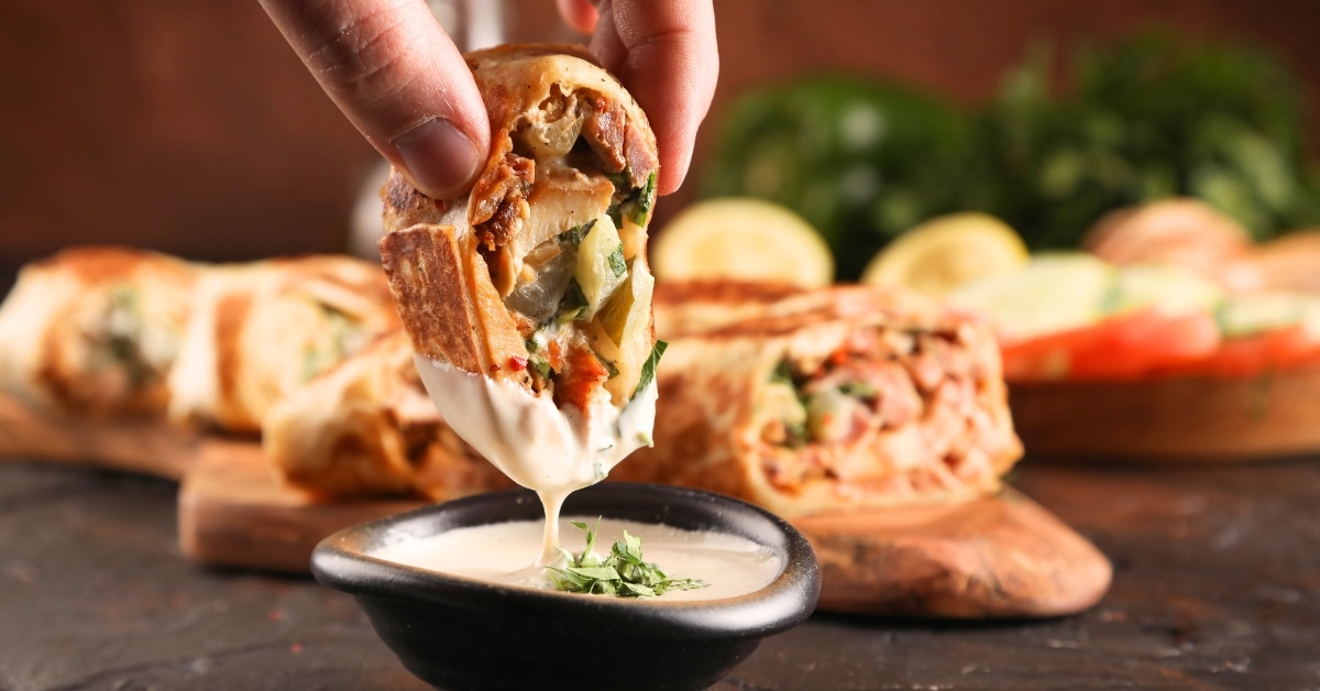 A person dipping a piece of shawarma into a small bowl of creamy sauce