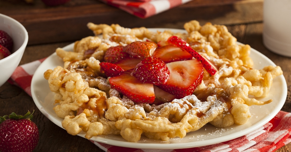 Funnel cake on a white plate with slices of strawberries and powdered sugar.