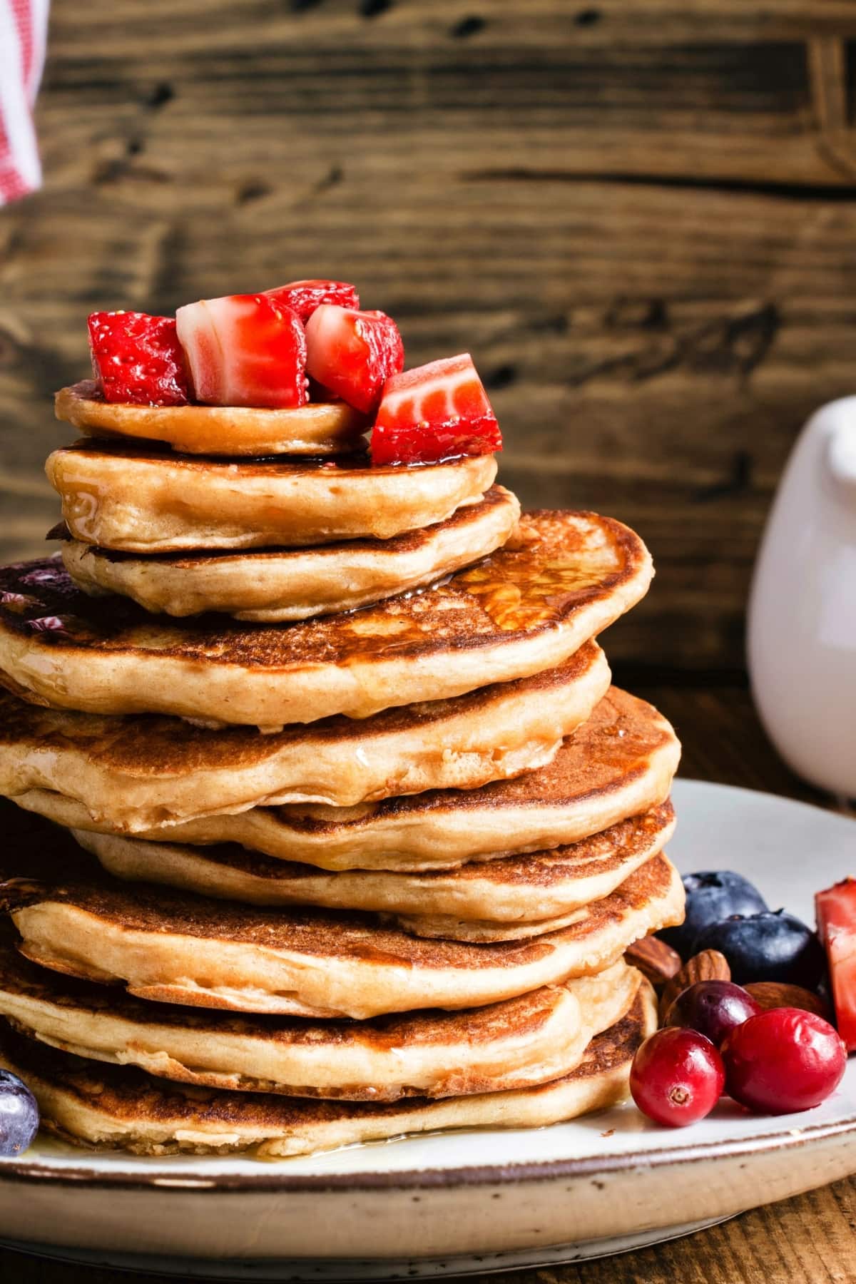 Pancakes stacked on a white plate topped with sliced strawberry garnished and dripping maple syrup.