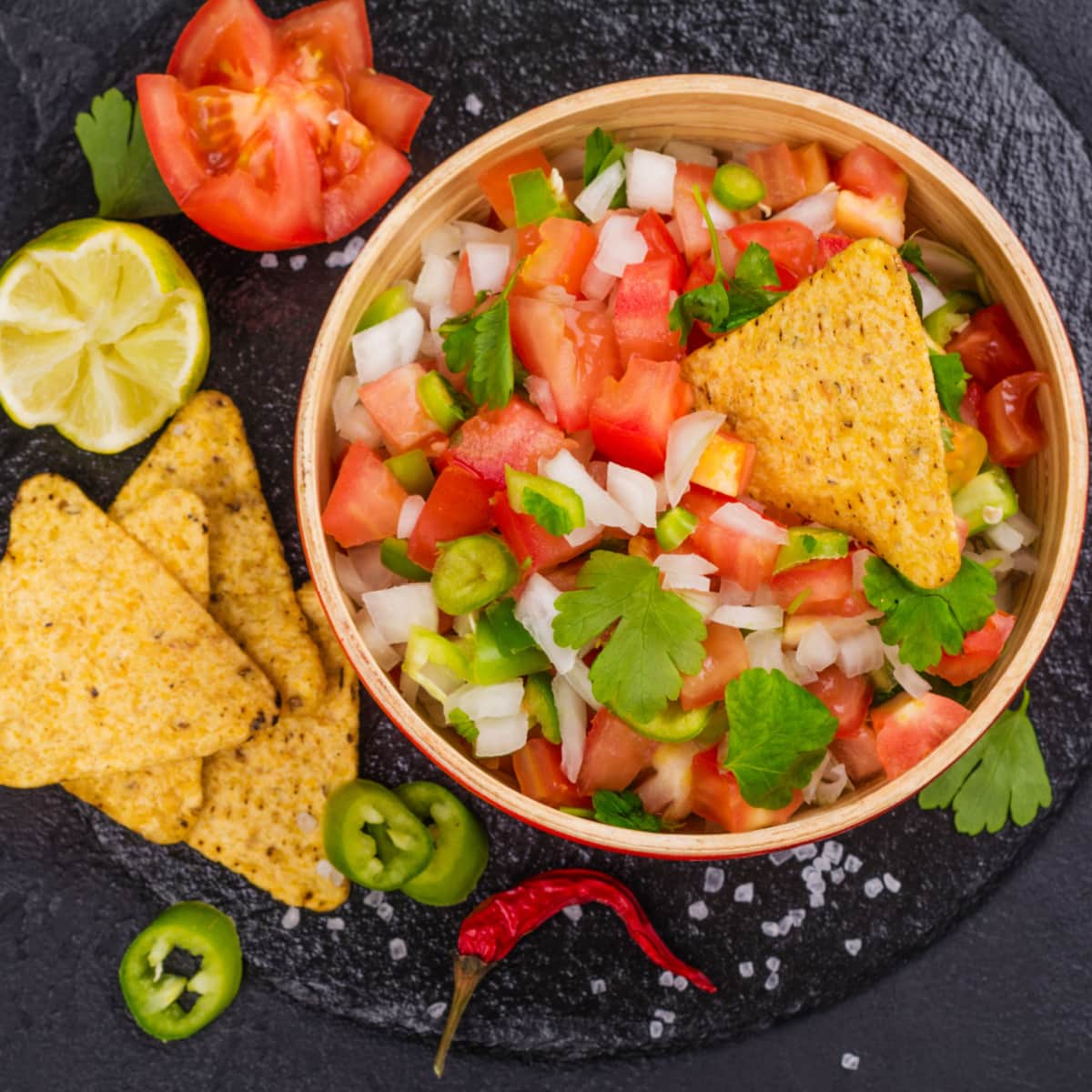 Nachos dipped on diced tomatoes, onions, cilantro, and jalapenos in red bowl. 