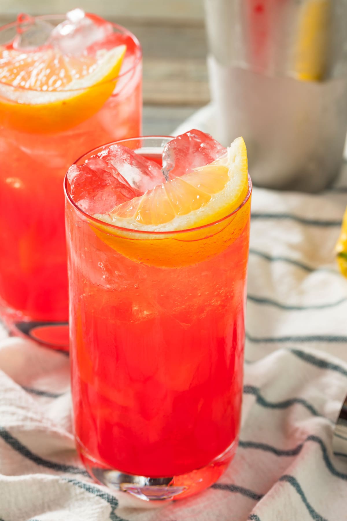 Orangey peach colored cocktail on ice served on a tall glass garnished with lemon wedge.