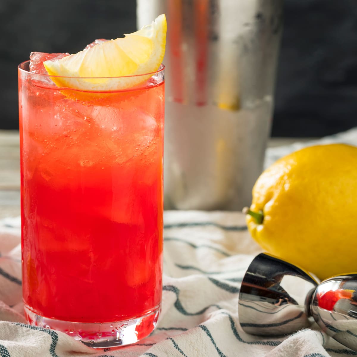 Sloe fizz gin cocktail in a tall glass filled with ice garnished with lemon slice, beside shaker and jigger on a table. 