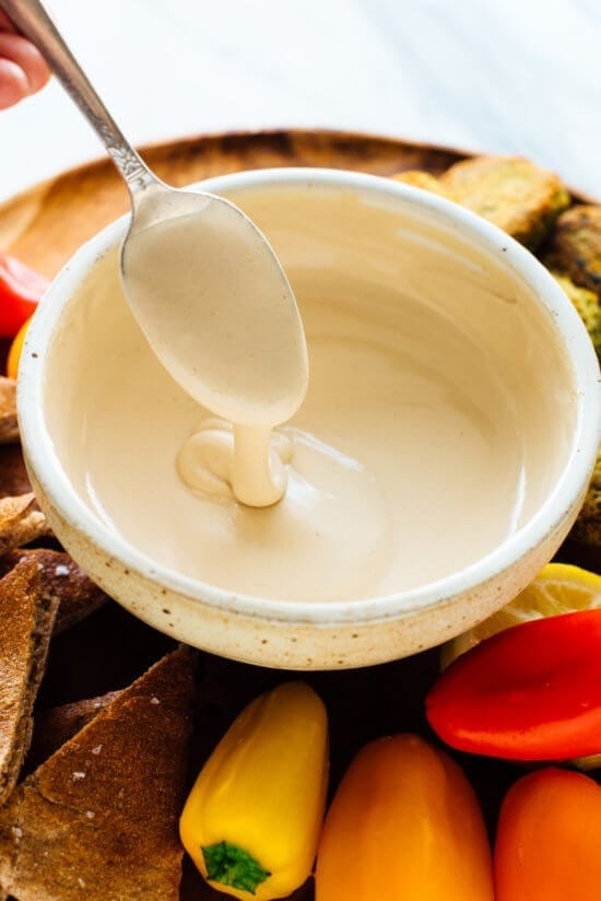 Spoon dripping with tahini sauce from a bowl served in a vegetable and bread platter.