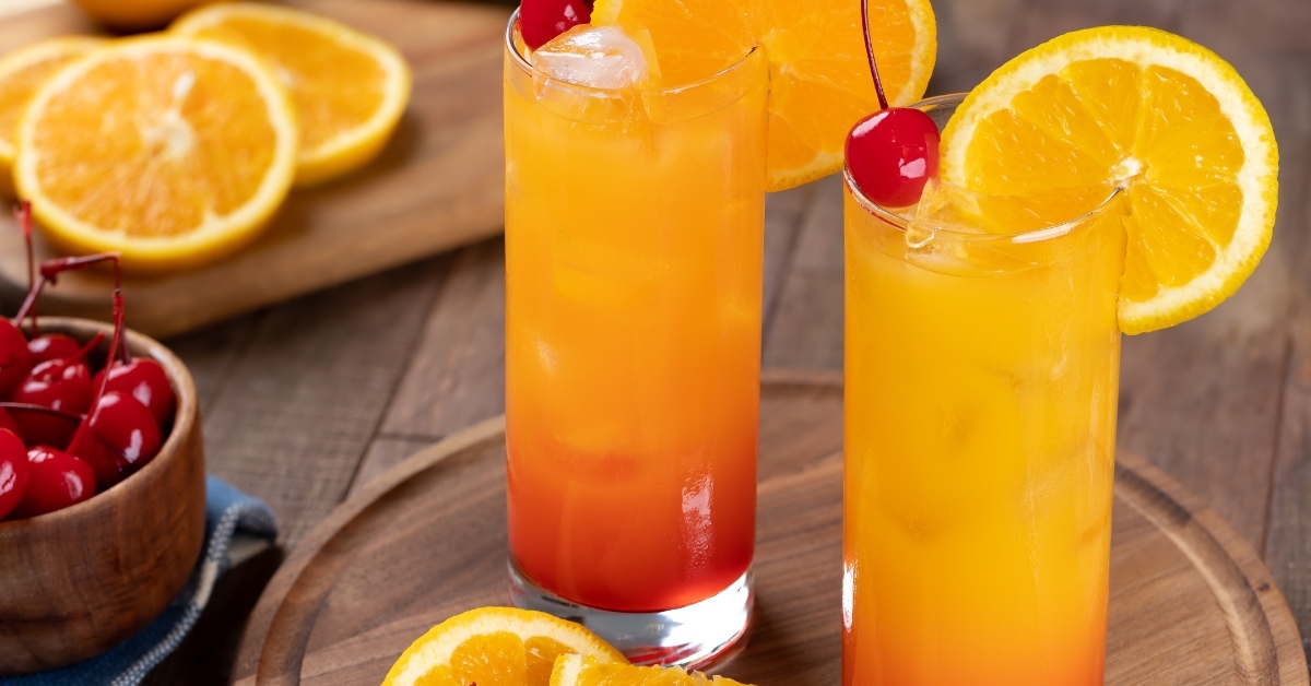 Two glasses of sunrise cocktail on a wooden tray served with orange slices and red cherries garnish.