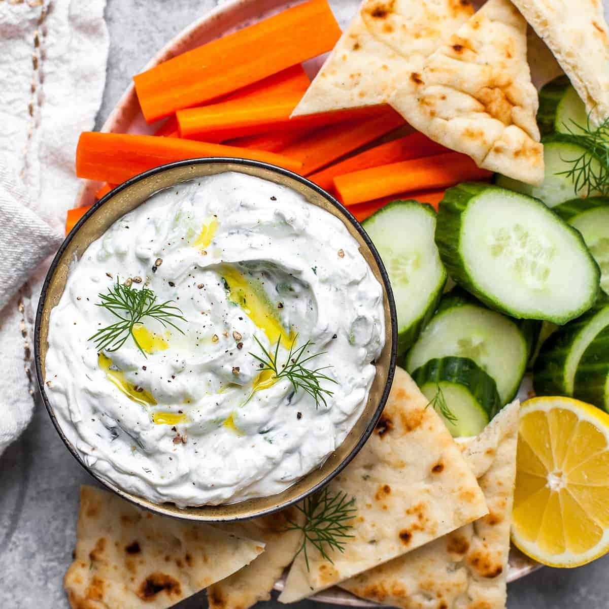 Tzatsiki sauce on a wooden bowl served with sliced cucumber, carrots, and pita bread.