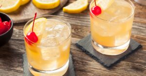 Two glasses of whiskey sour cocktail on top of a wooden table garnished with lemon and cherry.