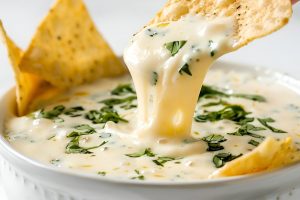 Dipping Chip into Mexican White Queso Cheese Dip in a White Bowl with Satisfying Cheese Pull