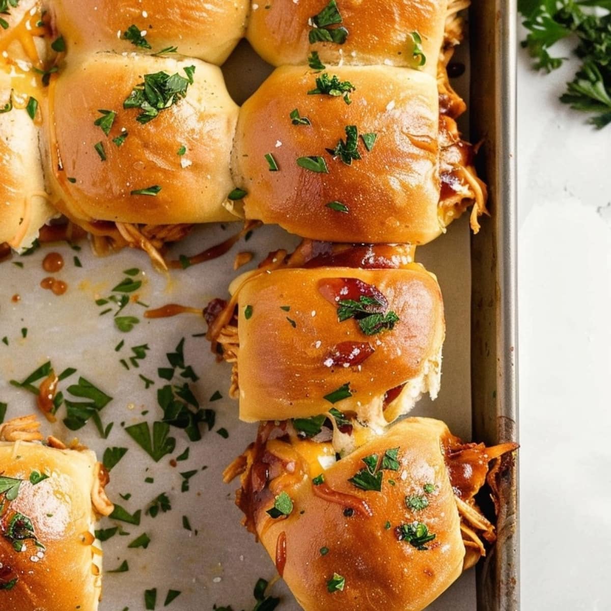 Pull apart slider rolls with shredded chicken bbq, cooked red onion slices, melted white cheese, and dill pickles filling dripping with sauce on a wooden board on a white marble table