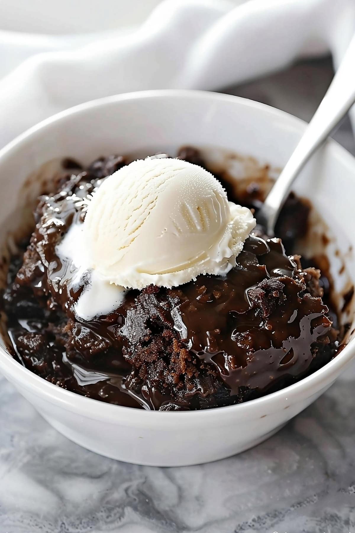 Chocolate cobbler in a white bowl garnished with scoop of vanilla ice cream with soon on the side.
