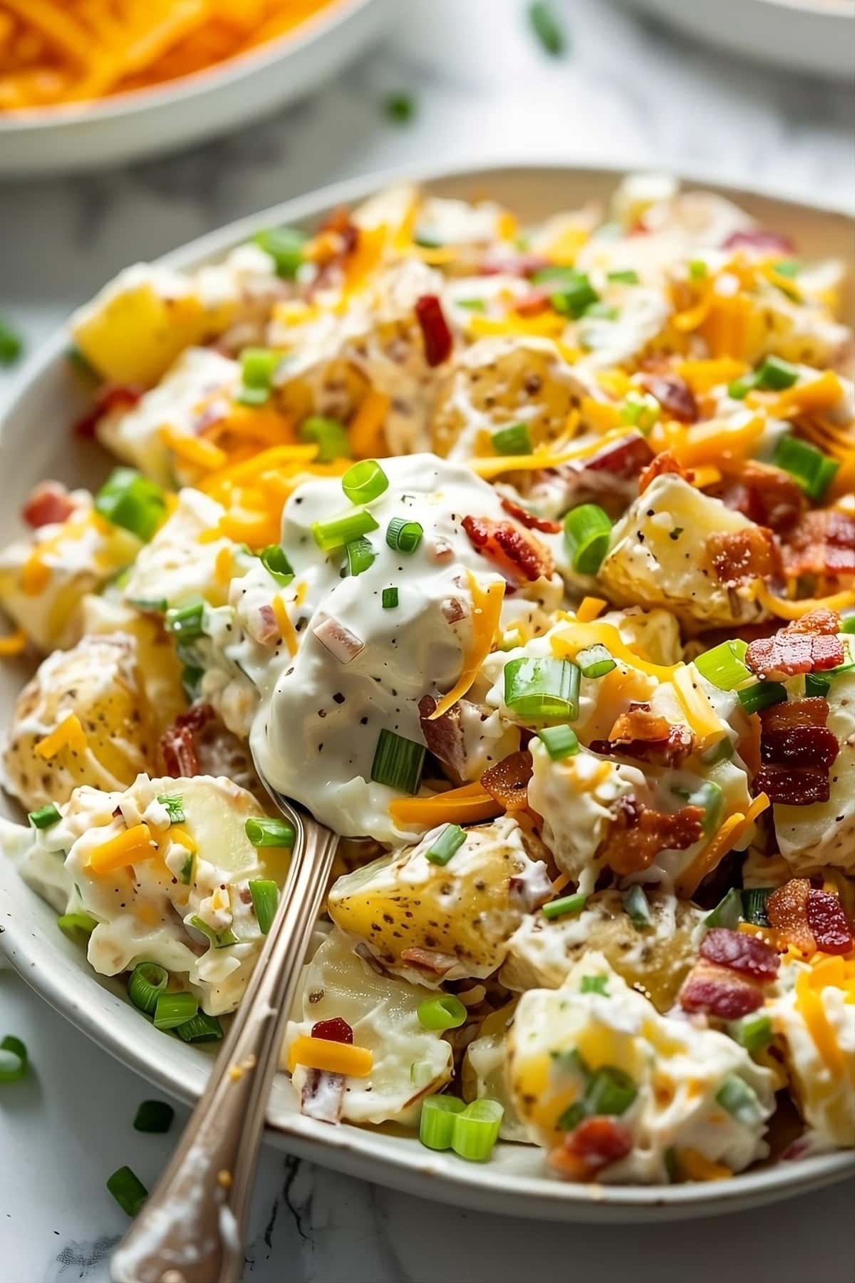 Loaded baked potato salad topped with lots of shredded cheese, bacon and green onions in a white bowl with spoon.