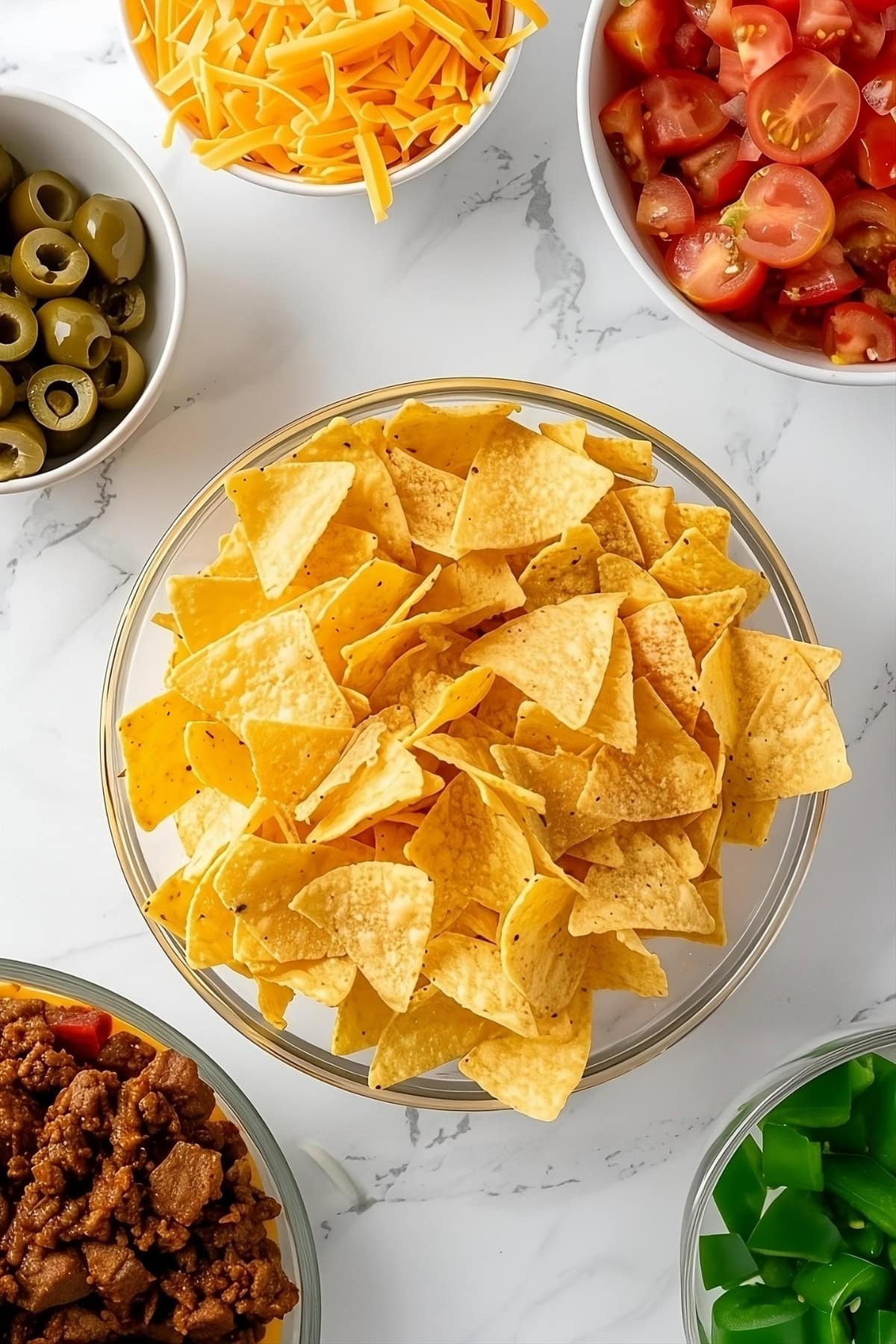 Tomatoes, grated cheese, tortilla chips, olives, beef and bell pepper on a table, top down view.