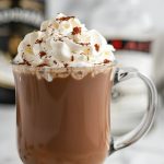 Bailey's Hot Chocolate in a Glass Mug with Whipped Cream and Cocoa Powder with Bottles of Bailey's in the Background
